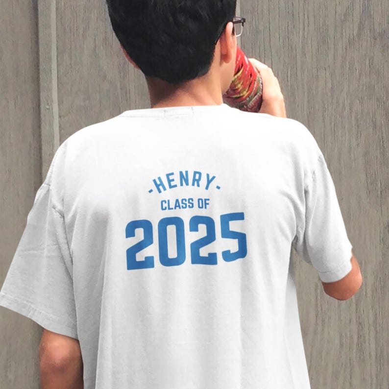May include: A light gray t-shirt with the name "HENRY" and "CLASS OF 2025" printed in blue. The person is holding a red and yellow object. The shirt is a crew neck style.