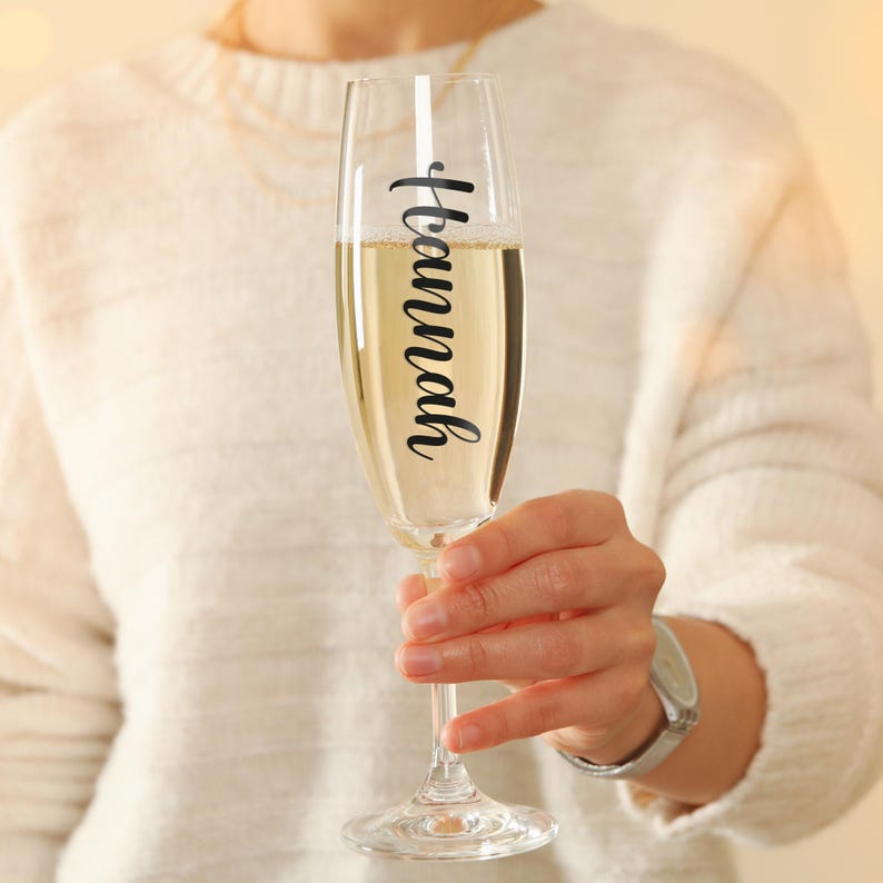May include: A clear glass champagne flute with the name "Hannah" written on it in black lettering. The flute is filled with champagne and is being held by a person.