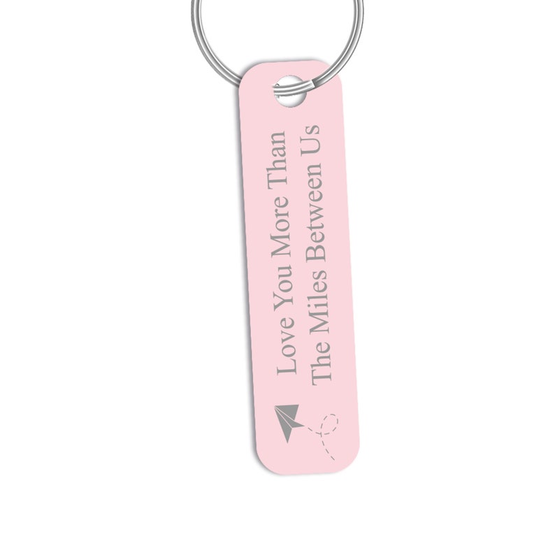 Puede incluir: Llavero rosa con un anillo plateado y el texto "I Love You More Than The Miles Between Us" con un avi&oacute;n de papel.