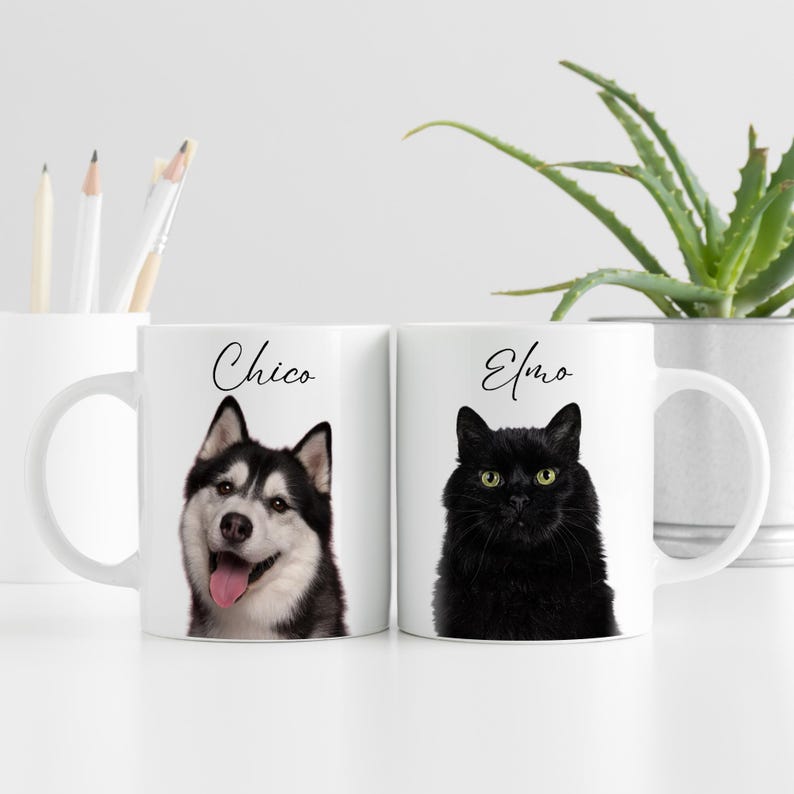 May include: Two white ceramic mugs with pet portraits. One mug features a black and white husky with the name "Chico" in script. The other mug displays a black cat with the name "Elmo" in script. A plant and art supplies are in the background.