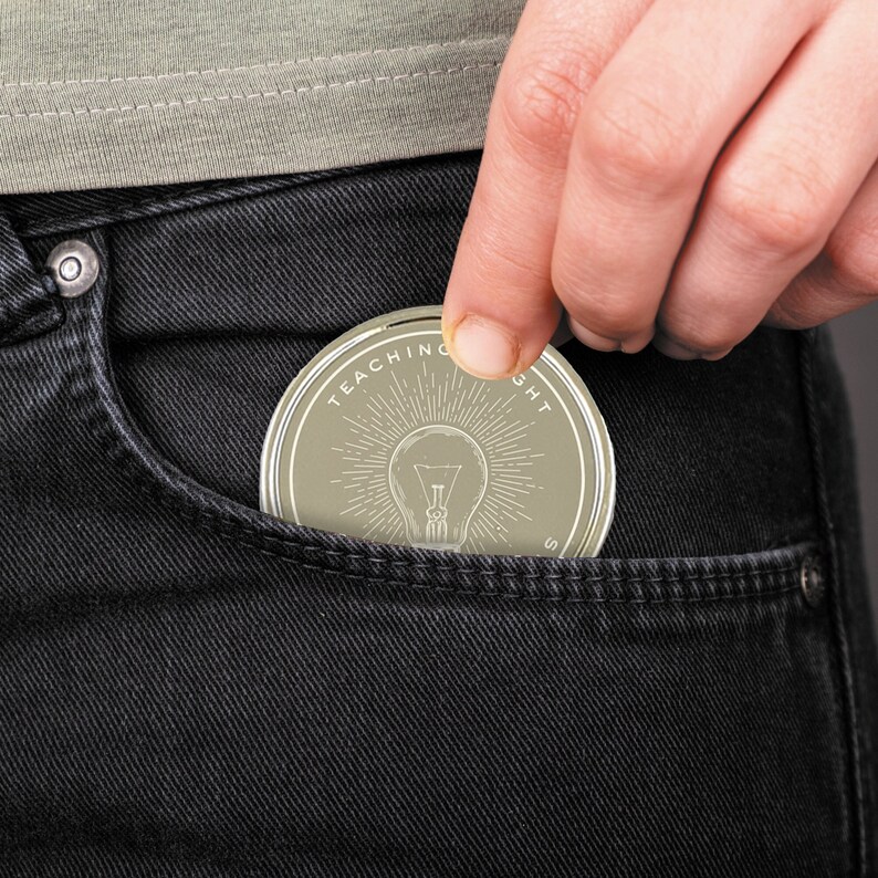 Puede incluir: Una moneda redonda y clara con las palabras "TEACHING THOUGHT" que rodean un dise&ntilde;o de bombilla. La moneda se est&aacute; metiendo en el bolsillo de unos vaqueros negros. La bombilla tiene l&iacute;neas radiantes.