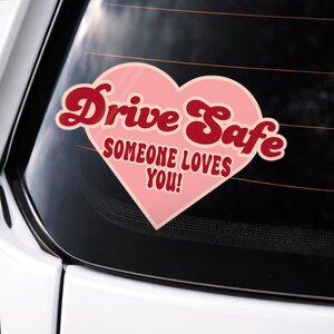 May include: A pink heart-shaped car sticker with the words "Drive Safe" in a retro font, and the message "Someone Loves You!" in a smaller font. The sticker is on a car window.