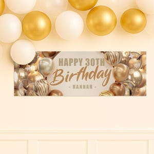 May include: A banner with the text "HAPPY 30TH Birthday - Hannah -" in gold script, surrounded by gold, silver, and white balloons. The banner is rectangular and set against a cream background, with additional balloons above.