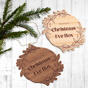 以下が含まれることがあります： 花柄のデザインと「Olivia's Christmas Eve Box」と「Annabelle's Christmas Eve Box」の文字が書かれた木製のクリスマスイブボックスのオーナメント2つ。