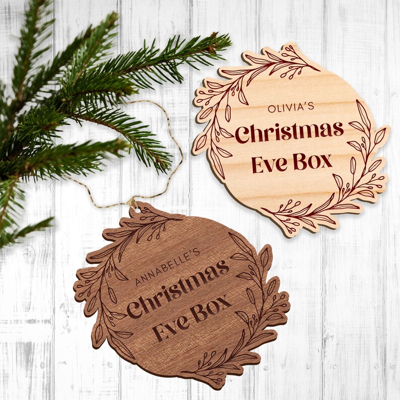 以下が含まれることがあります： 花柄のデザインと「Olivia's Christmas Eve Box」と「Annabelle's Christmas Eve Box」の文字が書かれた木製のクリスマスイブボックスのオーナメント2つ。