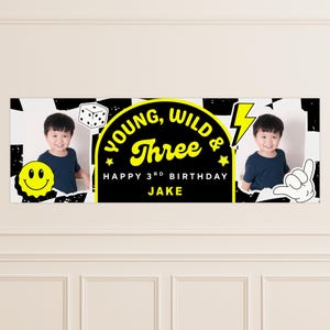 May include: A birthday banner with a black and white checkered background, featuring a photo of a child on each side. The banner has the text "Young, Wild & Three" in yellow, with "Happy 3rd Birthday Jake" below.