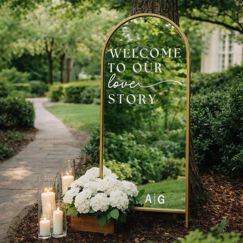 May include: A gold-framed sign with the words "WELCOME TO OUR love STORY" in white script. The sign is placed outdoors near a path, with white flowers in a wooden box and candles in glass holders at the base. The initials "A G" are at the bottom.