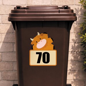 May include: A brown trash bin with a brown lid. A sticker of a cartoon Highland cow is on the bin, with the number 70 on a rectangular sign below the cow.