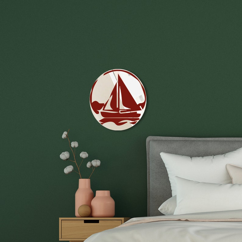 May include: A red sailboat silhouette on a white background in a round mirror. The mirror is hanging on a dark green wall above a wooden nightstand with a vase of white cotton flowers.