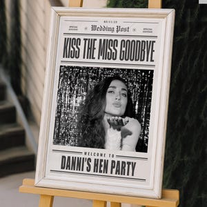May include: A framed sign with the text "KISS THE MISS GOODBYE" and "DANNI'S HEN PARTY". The black and white image inside the frame shows a woman blowing a kiss. The frame is white and sits on a wooden easel.