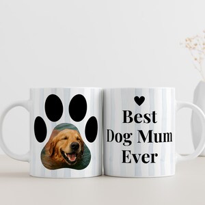 May include: White ceramic mug with a paw print design featuring a photo of a golden retriever. The other side of the mug has the text "Best Dog Mum Ever" with a black heart. The mug has a blue and white striped background.