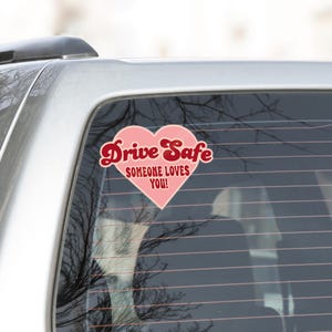 May include: A pink heart-shaped car sticker with the words "Drive Safe" in a cursive font and "SOMEONE LOVES YOU!" in a smaller font. The sticker is on a car window.