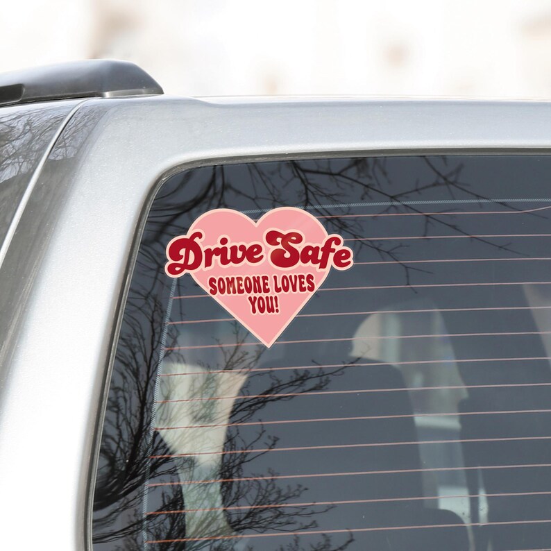 May include: A pink heart-shaped car sticker with the words "Drive Safe" in a cursive font and "SOMEONE LOVES YOU!" in a smaller font. The sticker is on a car window.