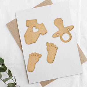 May include: Wooden cutouts of a baby onesie, a pacifier, and two baby feet. The onesie has a small heart cutout in the center.