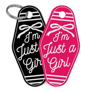 K&ouml;nnte beinhalten: Zwei Schl&uuml;sselanh&auml;nger im Motel-Stil, einer schwarz und einer pink, mit dem Schriftzug "I'm Just a Girl" in wei&szlig;er Schrift. Jeder Schl&uuml;sselanh&auml;nger hat oben ein Schleifendesign und einen silbernen Schl&uuml;sselring.