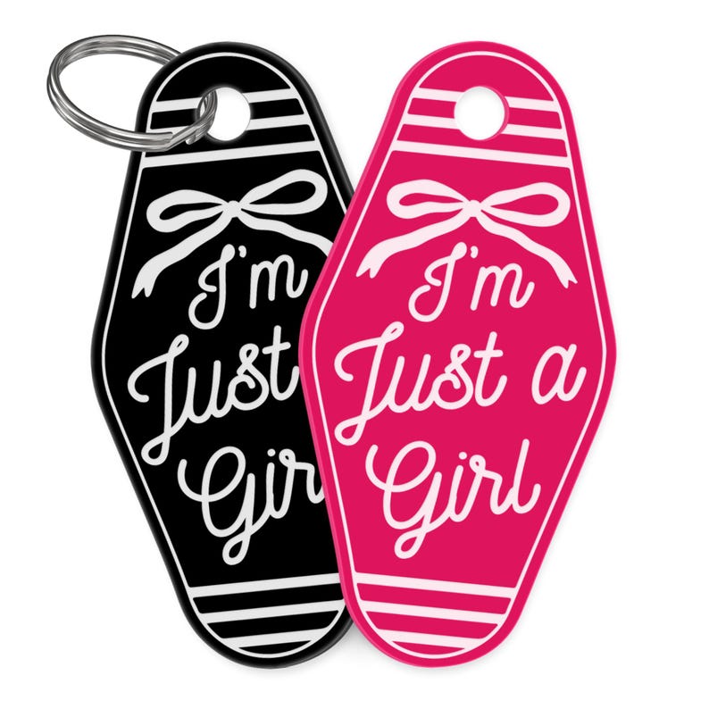 K&ouml;nnte beinhalten: Zwei Schl&uuml;sselanh&auml;nger im Motel-Stil, einer schwarz und einer pink, mit dem Schriftzug "I'm Just a Girl" in wei&szlig;er Schrift. Jeder Schl&uuml;sselanh&auml;nger hat oben ein Schleifendesign und einen silbernen Schl&uuml;sselring.