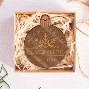 May include: A wooden ornament shaped like a Christmas tree bauble with the text "Promoted to Grandparents Est. 2023" engraved on it. The ornament is in a small brown box with shredded paper.