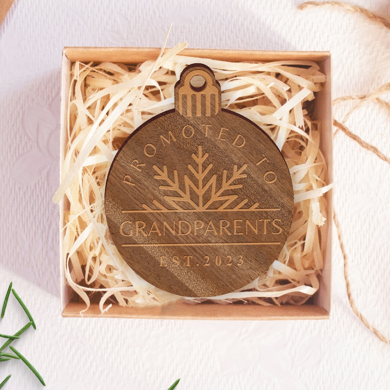 May include: A wooden ornament shaped like a Christmas tree bauble with the text "Promoted to Grandparents Est. 2023" engraved on it. The ornament is in a small brown box with shredded paper.