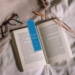May include: A blue bookmark with the text "book babe" and a small flower design. The bookmark is placed on an open book with text in Dutch.