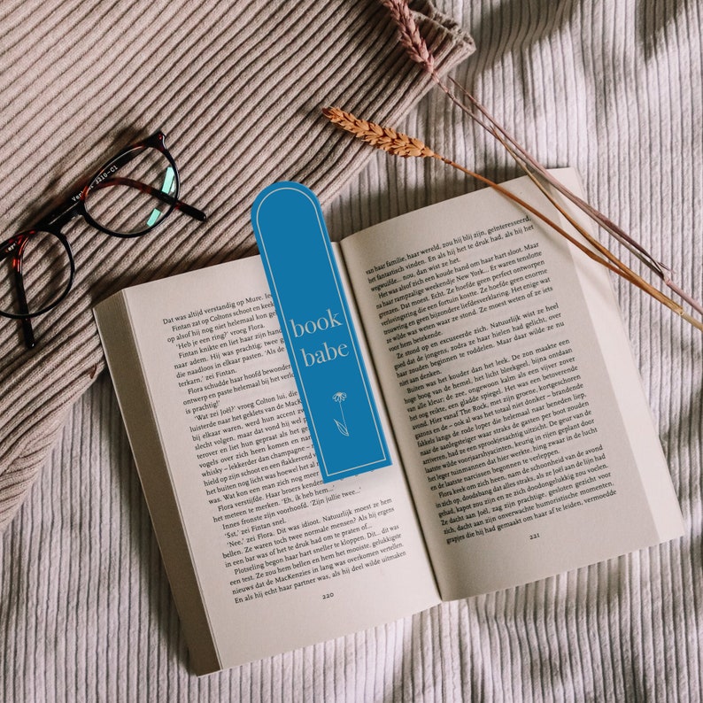 May include: A blue bookmark with the text "book babe" and a small flower design. The bookmark is placed on an open book with text in Dutch.