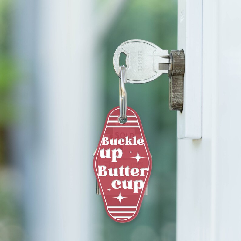 May include: Red motel key tag with white text that reads "Buckle up Butter Cup" with two white stars on either side of the text. The key tag is attached to a silver key.