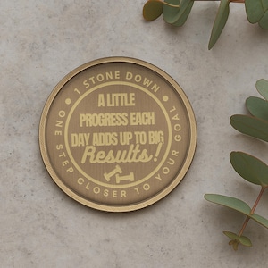 Puede incluir: Un token redondo de color dorado con el texto "One Step Closer To Your Goal" y "A Little Progress Each Day Adds Up To Big Results!" El token tiene un borde texturizado.