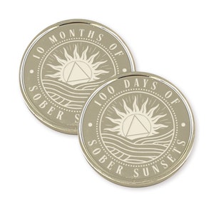 May include: Two silver-colored coins with a beige background. The coins feature a sun with a triangle inside, waves, and the text "100 Days of Sober Sunsets" and "100 Months of Sober Sunsets".