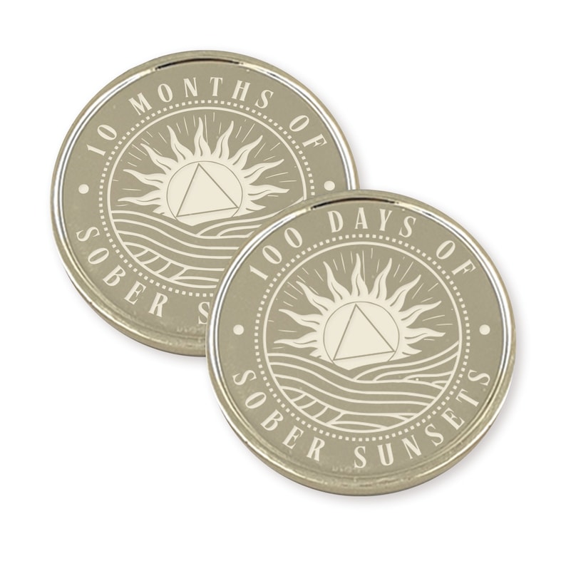 May include: Two silver-colored coins with a beige background. The coins feature a sun with a triangle inside, waves, and the text "100 Days of Sober Sunsets" and "100 Months of Sober Sunsets".