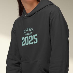 May include: Black hoodie with the name "Rachel" and "Class of 2025" printed in a light blue, textured font. The hoodie has a drawstring hood and a front pocket. A graduation-themed apparel item.
