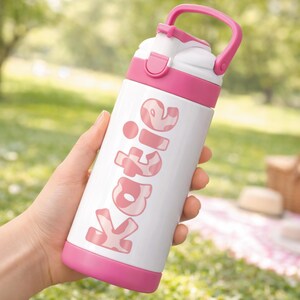 May include: A white and pink water bottle with the name "Katie" printed on the side. The bottle has a pink handle, lid, and base. The background is a blurred outdoor scene with green grass.