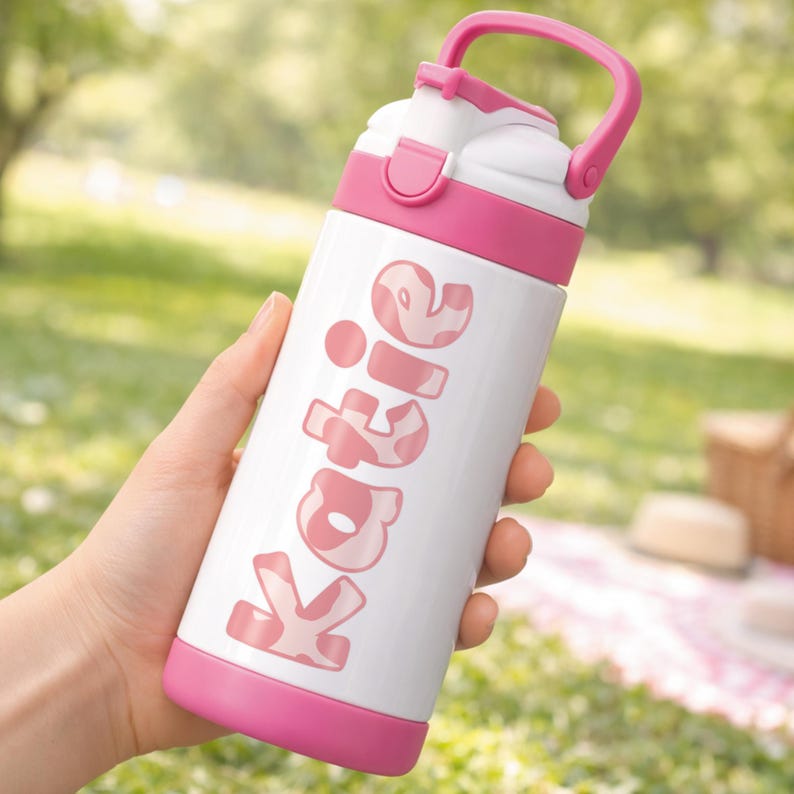 May include: A white and pink water bottle with the name "Katie" printed on the side. The bottle has a pink handle, lid, and base. The background is a blurred outdoor scene with green grass.
