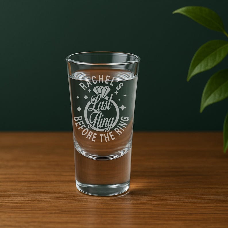 May include: A clear shot glass filled with liquid, with the text "Rachel's Last Fling Before The Ring". The glass is on a wooden surface with a dark green background and a leafy plant.