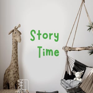 May include: A white wall with the words "Story Time" written in green letters. A brown woven giraffe toy stands on the left side of the image. A hanging hammock chair with a gray cushion and a white stuffed reindeer toy is on the right side of the image.