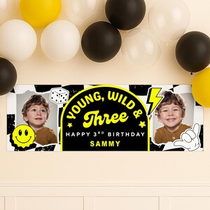 May include: A personalized birthday banner with the text "Young, Wild & Three" in yellow on a black background. The banner includes a photo of a child, a smiley face, and a dice graphic. Balloons in yellow, white, and black are in the background.