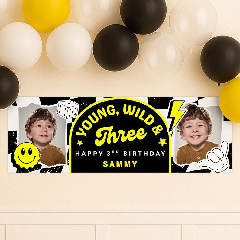 May include: A personalized birthday banner with the text "Young, Wild & Three" in yellow on a black background. The banner includes a photo of a child, a smiley face, and a dice graphic. Balloons in yellow, white, and black are in the background.