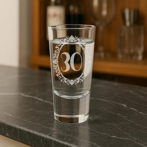 May include: A clear shot glass filled with water, featuring the number "30" in a decorative white floral frame. The glass is set on a dark gray surface, with a blurred background of bottles and glassware.