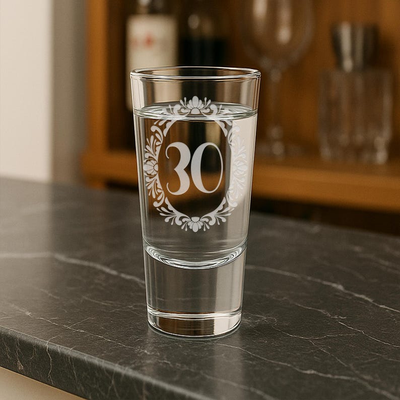 May include: A clear shot glass filled with water, featuring the number "30" in a decorative white floral frame. The glass is set on a dark gray surface, with a blurred background of bottles and glassware.
