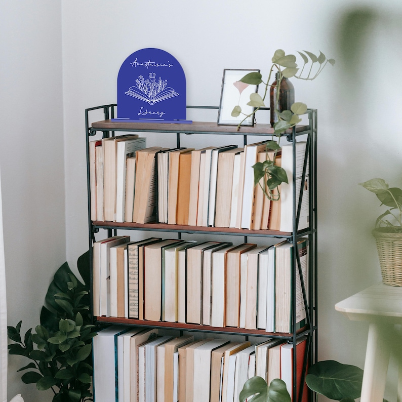 May include: A black metal bookshelf with three shelves filled with books. A blue bookend with a floral design and the text "Anastasia's Library" is on the top shelf. A small framed picture is on the shelf next to the bookend.