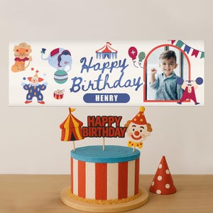 Puede incluir: Una escena festiva de cumplea&ntilde;os con una tarta tem&aacute;tica de circo con rayas rojas y blancas, una parte superior azul y adornos decorativos. Una pancarta encima muestra ilustraciones de circo, las palabras "Happy Birthday Henry" y una foto de un ni&ntilde;o.