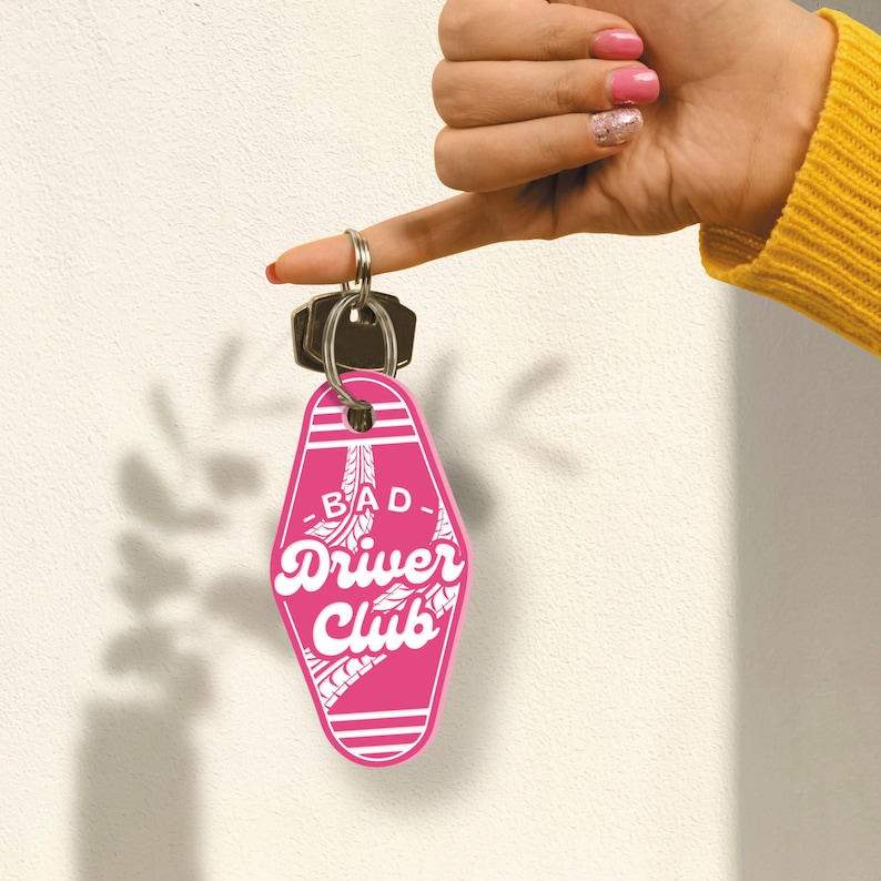 May include: Pink motel key chain with white text that reads "Bad Driver Club".