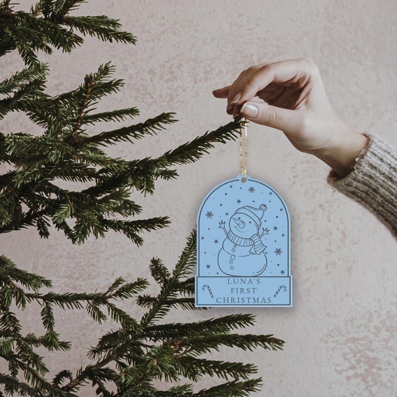 May include: A light blue Christmas ornament with a snowman design and the text "Luna's First Christmas".