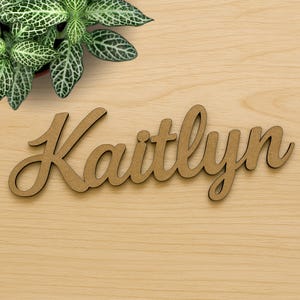 May include: A wooden sign with the name "Kaitlynn" written in cursive script. The sign is a light brown color and is made of wood.