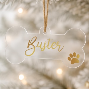 以下が含まれることがあります： 「Buster」の金色の筆記体と金色の足跡が特徴の、透明なアクリル製の犬の骨型オーナメント。 天然の麻ひもで吊り下げられており、ペット愛好家やホリデーデコレーションに最適です。