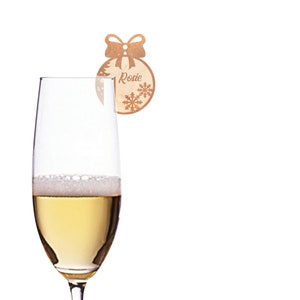 May include: A wooden Christmas ornament with a bow and snowflakes. The ornament has the name "Rosie" written on it. The ornament is hanging from a champagne flute.