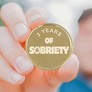 Pu&ograve; includere: Una moneta d'oro con il testo "3 Years of Sobriety" e un simbolo triangolare.