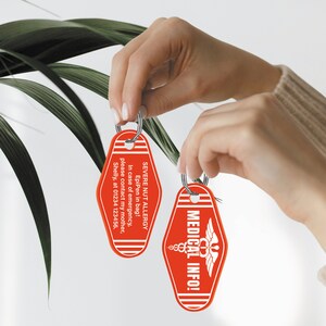 Puede incluir: Dos llaveros de motel rojos con texto blanco. Un llavero dice "SEVERE NUT ALLERGY EpiPen in bag! In case of emergency, please contact my mother, Shelly, at 01234 123456." El otro llavero dice "MEDICAL INFO!" con un s&iacute;mbolo m&eacute;dico.