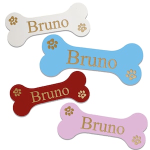 May include: Four wooden dog bone shaped tags with the name "Bruno" engraved on them. The tags are in white, light blue, red, and pink.