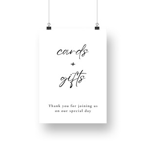 May include: A white sign featuring the words "cards + gifts" in black cursive script. The text "Thank you for joining us on our special day" is below. The sign is held by two black clips.