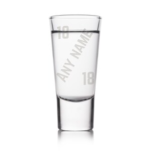 May include: A clear shot glass with the text "Any Name" and the number "18" etched on the side. The glass is filled with a clear liquid. The top of the glass has the number "18" etched on it.