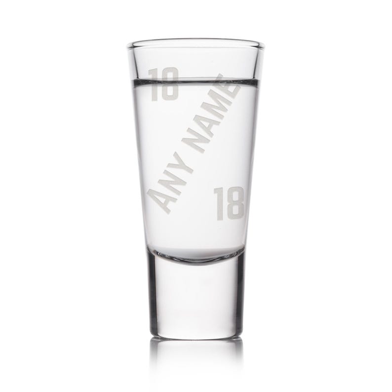 May include: A clear shot glass with the text "Any Name" and the number "18" etched on the side. The glass is filled with a clear liquid. The top of the glass has the number "18" etched on it.